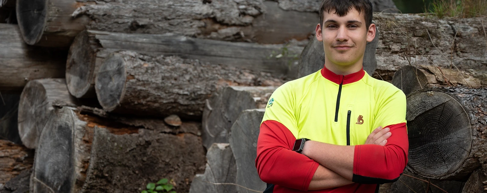 Foto von einem jungen Mann, der im Wald an einem Holzstapel lehnt.