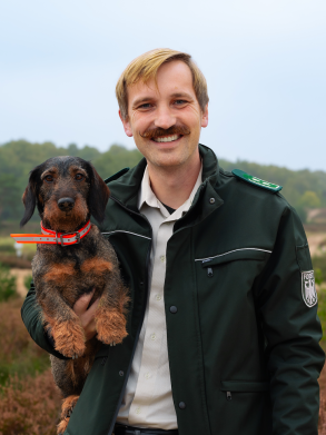 Ein Mann h&auml;lt einen Dackel unter seinem Arm. Er tr&auml;gt eine Bundesforst-Uniform und steht in der Heide.